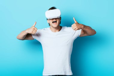 VR kulaklıklı genç adam mavi arka planda baş parmak gösteriyor.