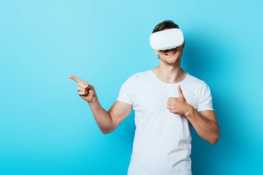 Beyaz tişörtlü ve VR kulaklı bir adam baş parmağını yukarı kaldırıyor ve mavi arka planda işaret ediyor.