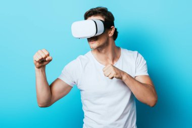 Beyaz tişörtlü ve vr kulaklı bir adam mavi arka planda yumrukları gösteriyor.