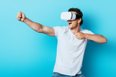 Mavi arka planda vr kulaklık kullanırken beyaz tişörtlü genç adam el kol hareketi yapıyor