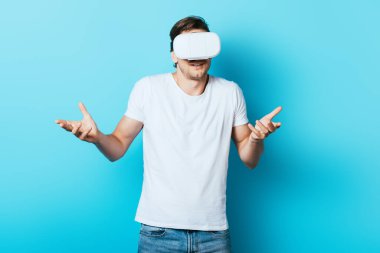 Beyaz tişörtlü ve VR kulaklı bir adam mavi arka planda omuz silkme hareketi gösteriyor.