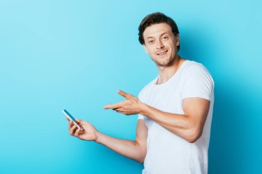 Beyaz tişörtlü genç adam mavi arka plandaki akıllı telefonu işaret ediyor.