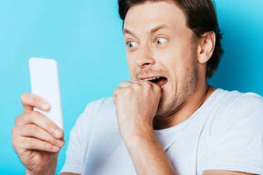 Selective focus of scared man using smartphone on blue background clipart
