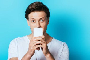 Genç adam mavi arka planda akıllı telefonla ağzını kapatıyor.