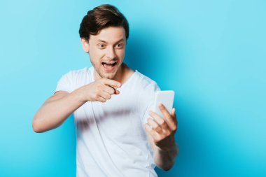Mavi arka plandaki akıllı telefonu işaret eden heyecanlı adamın seçici odağı