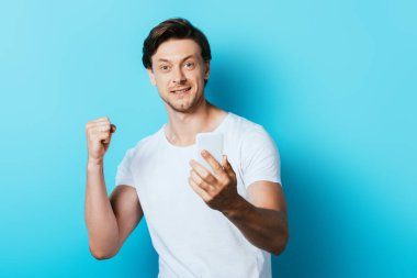 Genç adam kameraya bakıyor ve mavi arka planda akıllı telefonu tutarken evet hareketi gösteriyor.