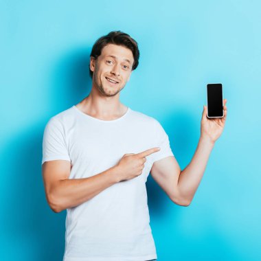 Beyaz tişörtlü genç adam mavi arka plandaki akıllı telefonu işaret ediyor.