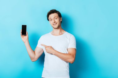 Beyaz tişörtlü adam mavi arka plandaki akıllı telefonu işaret ediyor.