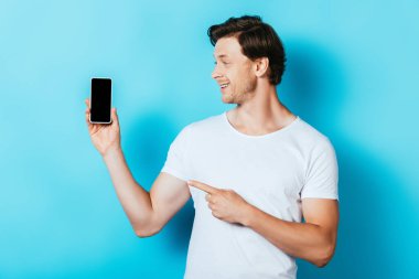 Adam mavi arka planda boş ekran ile akıllı telefonu işaret ediyor.