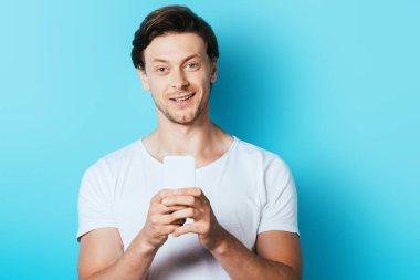 Genç adam mavi arka planda akıllı telefon kullanırken kameraya bakıyor.