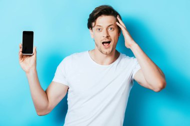 Heyecanlı adam boş ekranlı akıllı telefonu gösteriyor ve mavi arkaplandaki kameraya bakıyor.