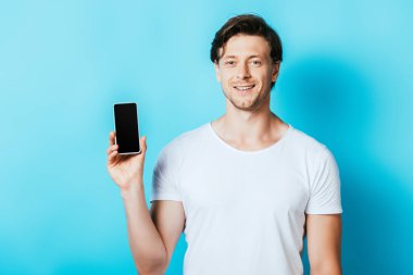 Genç adam mavi arka planda boş ekranlı akıllı telefonu gösterirken kameraya bakıyor