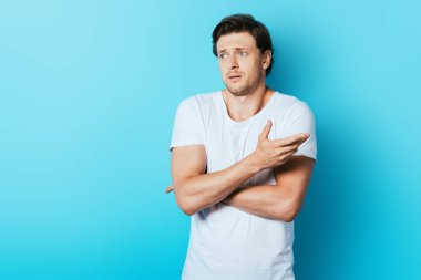 Kafası karışmış bir adam mavi arka plana bakarken elini işaret ediyor.