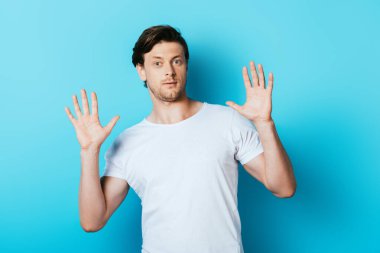 Beyaz tişörtlü kafası karışmış bir adam mavi arka plandaki kameraya bakıyor.