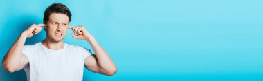 Panoramic shot of irritated man covering ears with fingers on blue background clipart