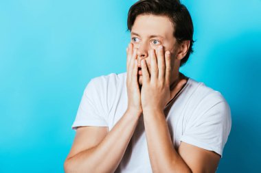 Shocked man with hands near cheeks looking away on blue background clipart