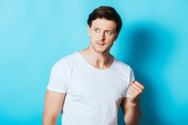 Beyaz tişörtlü kızgın adam mavi arka planda yumruğunu gösteriyor.