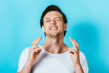Young man in white t-shirt with crossed fingers on blue background clipart