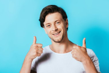 Beyaz tişörtlü genç adam mavi arka planda kameraya baş parmağını gösteriyor.