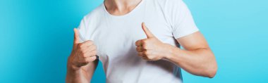 Beyaz tişörtlü bir adamın panoramik görüntüsü mavi arkaplanda başparmaklarını gösteriyor