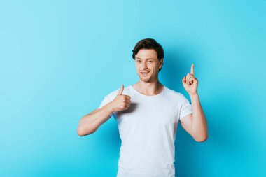 Young man in white t-shirt showing thumb up and having idea on blue background clipart