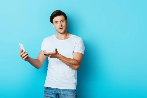 Homem Confuso Apontando Com Mão Para Smartphone Fundo Azul — Fotografia de Stock