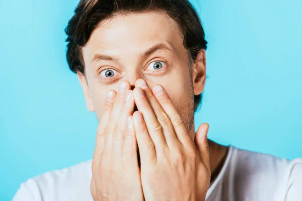 Shocked man looking at camera and covering mouth isolated on blue