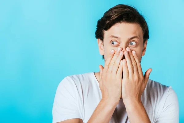 Surprised man covering mouth with hands isolated on blue