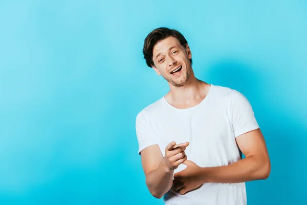 Smějící Muž Bílém Tričku Ukazuje Prstem Kameru Modrém Pozadí — Stock fotografie