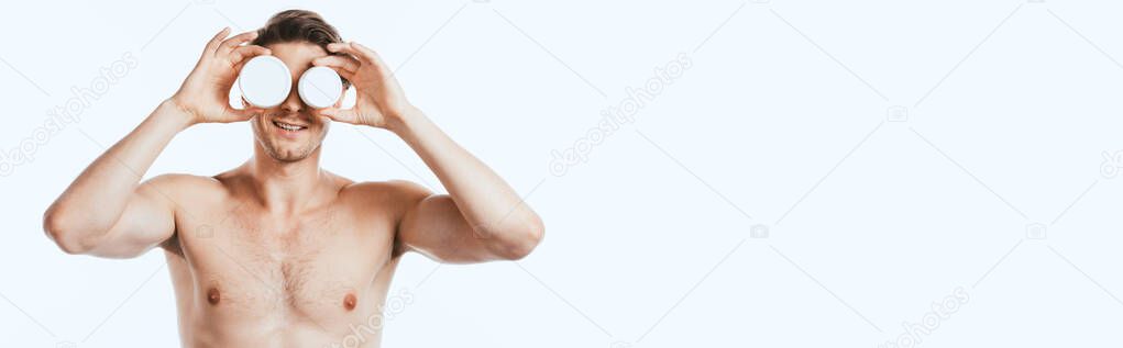 Panoramic crop of shirtless man holding jars of cosmetic creams near face isolated on white