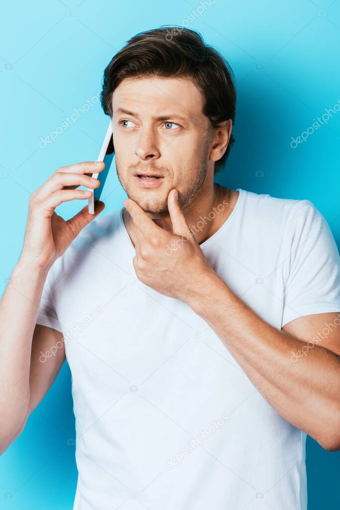 Pensive man touching chin while talking on smartphone on blue background