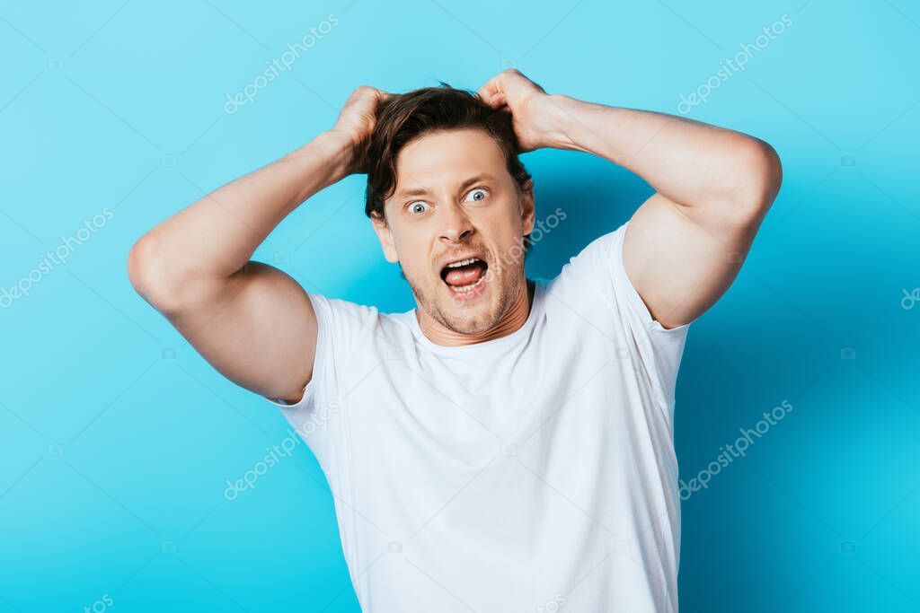 Crazy man with hands near hair in white t-shirt looking at camera on blue background