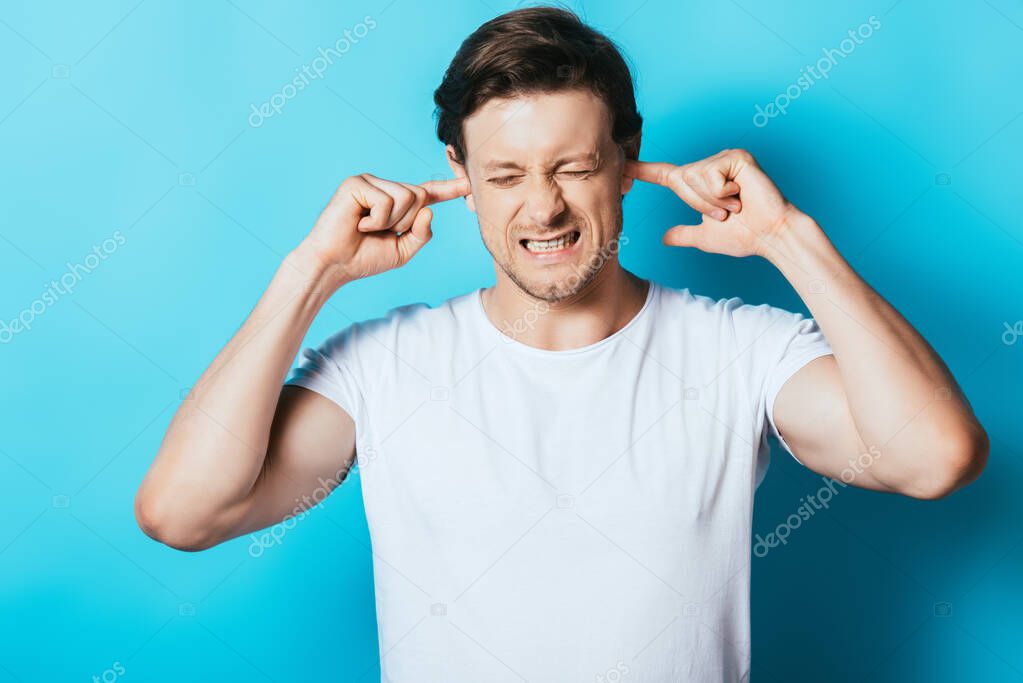 Angry man in white t-shirt covering ears with fingers on blue background