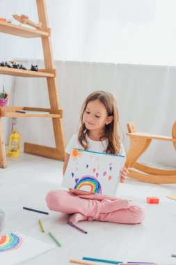 Pijamalı kız yerde oturuyor ve rengarenk keçeli kalemlerin yanında çizim yapıyor.
