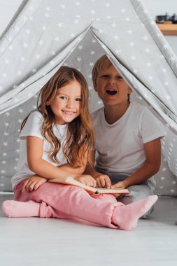excited brother and sister in pajamas looking at camera while sitting in kids wigwam and reading book clipart