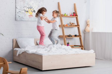 Erkek ve kız kardeşler pijamalarıyla yatağa atlarken el ele tutuşuyorlar.