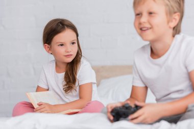 KYIV, UKRAINE - 21 Temmuz 2020: yatak odasında video oyunu oynayan heyecanlı erkek kardeşe bakan kitap seçici kız odaklı