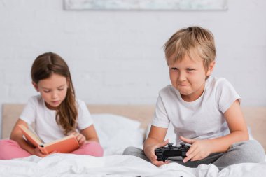 KYIV, UKRAINE - 21 Temmuz 2020: kız kardeşin yatak odasında kitap okuduğu yerde video oyunu oynayan konsantre çocuğun odak noktası