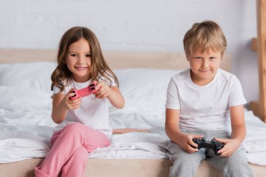 KYIV, UKRAINE - 21 Temmuz 2020: yatak odasında pijamalarıyla video oyunu oynayan heyecanlı kız ve odaklı kardeş