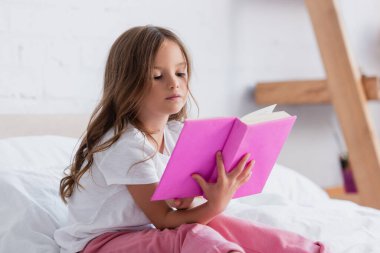 Pijamalarıyla yatakta kitap okuyan konsantre bir kız.