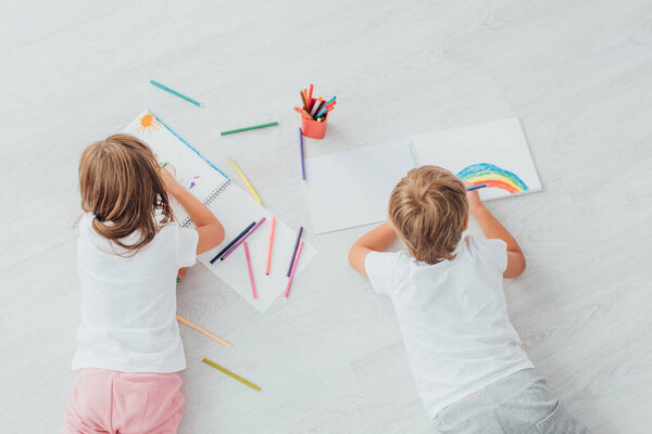 вид сверху на брата и сестру в пижаме лежащих на полу и рисунок с войлочными ручками в альбомах эскизов