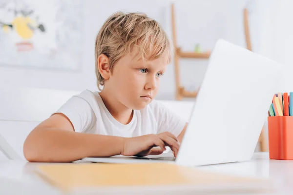 白いTシャツを着た少年がテーブルに座りノートパソコンを使って — ストック写真