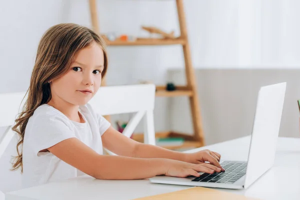 白いTシャツを着た女の子がテーブルに座ってノートパソコンを使いながらカメラを見て — ストック写真