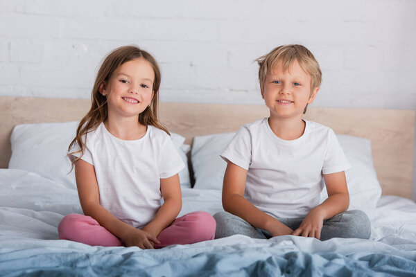 brother and sister in pajamas looking at camera while sitting with crossed legs on bed