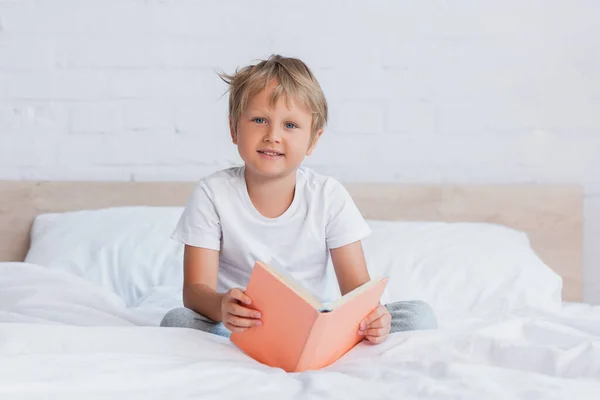 Chłopiec Białym Shirt Patrząc Aparat Podczas Siedzenia Łóżku Książki — Zdjęcie stockowe