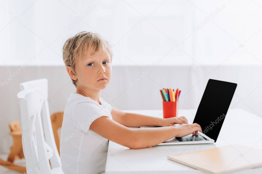 serious boy in white t-shirt looking at camera while sitting at table and using laptop