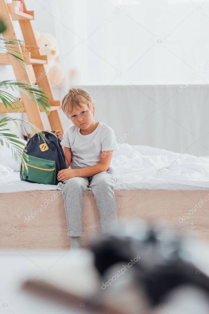 selective focus pf upset boy in pajamas looking at camera while sitting on bed near school backpack 