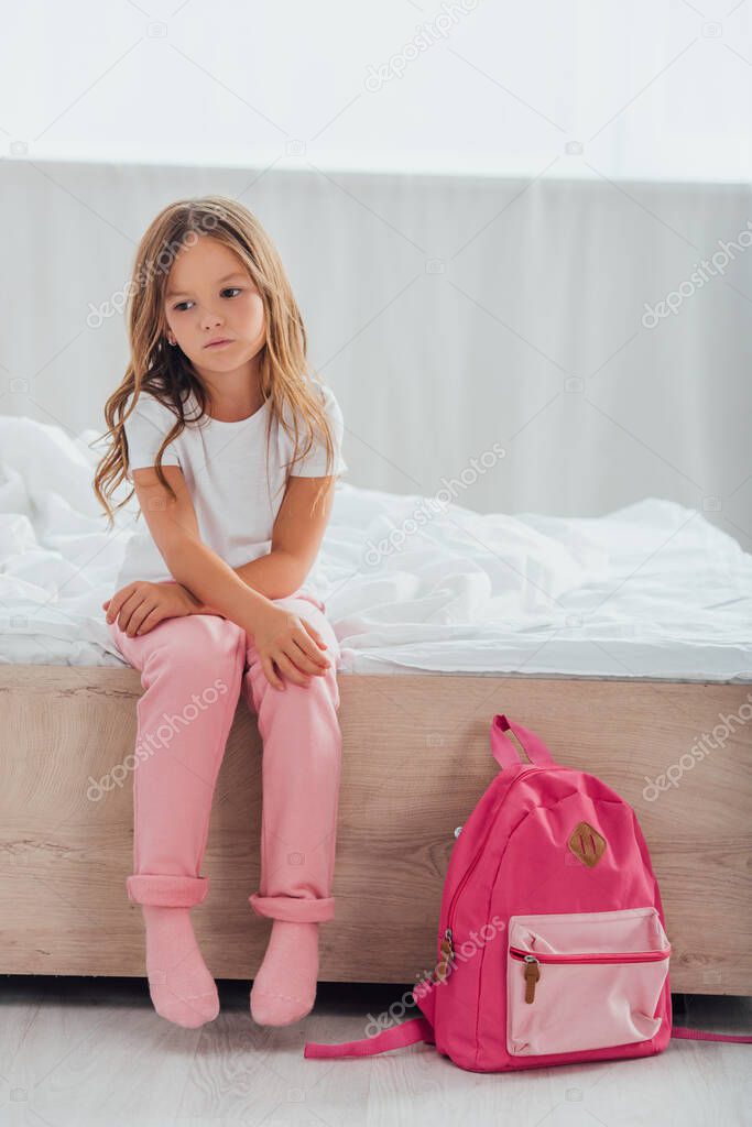 upset girl in pajamas sitting on bed near school backpack on floor