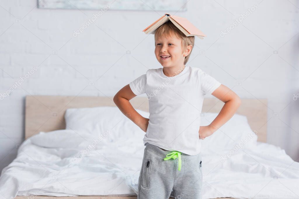 excited kid in white t-shirt standing with hands on hips and book on head in bedroom