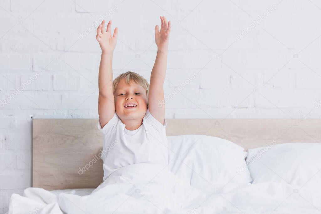 awakened boy in white t-shirt looking at camera while stretching in bed